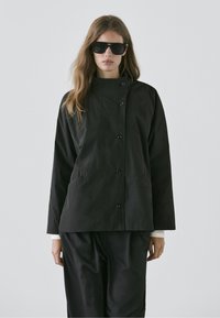 Vrouw met lang haar, gekleed in een zwart jasje met knopen, zwarte broek en grote donkere zonnebril, tegen een effen grijze achtergrond.