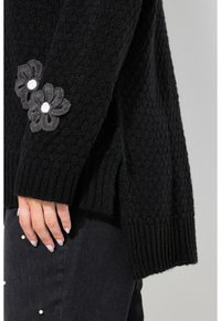 Mustat neuleet vastaavat kuvion ja ribattu helma, jossa on harmaita brodeerattuja kukkia valkoisilla keskiosilla vasemmassa hihassa.