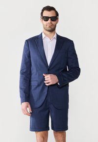 Blazer bleu marine assorti avec un short ajusté. Comprend un devant à deux boutons, des revers en pointe et une chemise blanche à boutons. Lunettes de soleil portées.