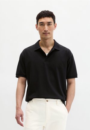 Man draagt een zwart poloshirt met korte mouwen, gestopt in een witte broek, staand tegen een effen witte achtergrond met een neutrale uitdrukking.