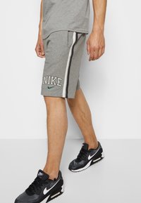 Muž v šedých Nike šortkách se postranními pruhy a černých teniskách Nike Air Max, stojící na jednobarevném bílém pozadí.