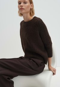 Dunkelbrauner Strickpullover mit strukturierter Oberfläche und weiter Passform, ausgestattet mit einem Rundhalsausschnitt und dreiviertellangen Ärmeln, kombiniert mit dunklen Stoffhosen.