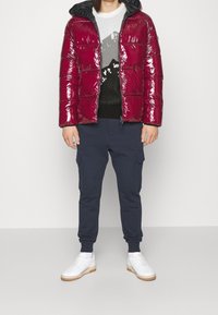 Giacca a vento rossa lucida con cappuccio, maglione nero con un design di montagna, joggers cargo blu navy e sneaker bianche con suola beige.