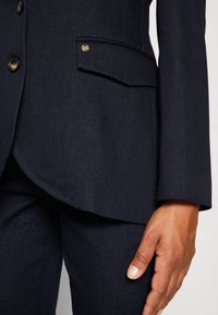 Tmavě modré sakové sako s texturovanou tkaninou, obsahující kovové knoflíky a přední kapsu. Detailní záběr na rukáv a umístění ruky.