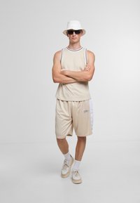 Beige Sportanzug bestehend aus einem ärmellosen Shirt mit Rundhalsausschnitt und passenden Shorts mit einem weißen Seitenpanel und Logo-Detail.