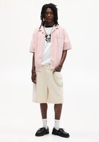 Camicia rosa chiaro a maniche corte sopra una maglietta bianca con grafica, pantaloni corti beige fino al ginocchio, scarpe nere e calzini bianchi con accenti blu.
