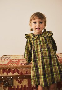 Braun-grün kariertes Kleid mit Rüschensaum und Puffärmeln, gefertigt aus leichtem Stoff, getragen von einem Kind neben einem gemusterten Koffer.