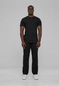 T-shirt noir à manches courtes associé à un pantalon noir. Les deux ont une texture lisse. Baskets blanches visibles. Le modèle se tient devant un fond gris.