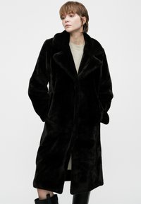Mulher vestindo um longo casaco de peluche preto sobre um vestido de cor clara, de pé com as mãos nos bolsos, olhando para o lado contra um fundo liso.