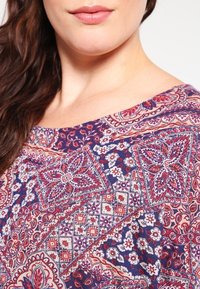 Blusa multicolor con un patrón floral y de paisley. Presenta un cuello redondo amplio y una tela ligera. Los colores predominantes son el azul marino, el rojo y el crema.