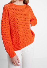 Femme portant un pull en tricot orange vif avec un motif texturé et un pantalon blanc, se tenant avec une main sur la hanche.