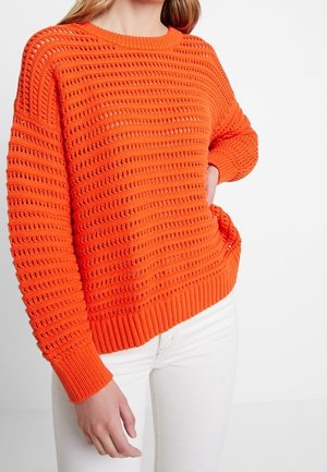 Vrouw in een feloranje gebreide trui met een structuurpatroon en witte broek, staat met één hand op de heup.