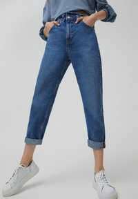 Jean en denim bleu taille haute avec revers roulés, présentant une coupe droite, cinq poches et des coutures contrastantes. Associé à des baskets blanches.