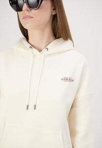 Krēmas krāsas džemperis ar priekšējo kabatu, piedāvā nelielu sarkanu un zilu "Guess Jeans" logo. Tas ir aprīkots ar sudraba krāsas pleķu vilkšanas auklām un ir brīvs piegriezums.