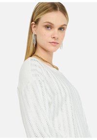 Maglione bianco con texture a righe verticali e una sottile lucentezza, caratterizzato da un ampio scollo e una vestibilità rilassata. Accentato da una collana di catene d'oro sovrapposte.