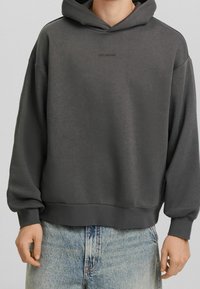 Felpa grigia realizzata in un morbido misto cotone, con una vestibilità comoda, polsini a coste e un piccolo logo sul petto. Abbinata a jeans azzurri chiari.