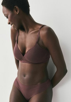 Femme portant un soutien-gorge et un slip mauve assortis, souriante et regardant vers le bas sur un fond clair uni.