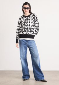 Juodas ir baltas megztinis su drąsiu pakartotiniu "DKNY" logotipu, derinamas su šviesiai mėlynomis plačiomis kelnėmis ir smailiais juodais batais.