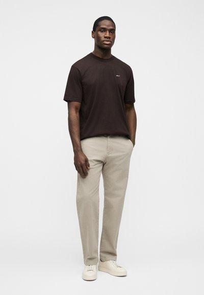 Άνδρας όρθιος με τα χέρια στις τσέπες, φοράει σκούρο καφέ κοντομάνικο μπλουζάκι, μπεζ παντελόνι και λευκά αθλητικά παπούτσια, σε φόντο απλό.