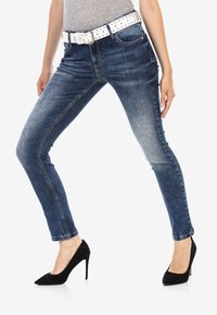 Jeans en denim bleu skinny avec une finition délavée et une ceinture cloutée blanche, associés à des chaussures à talons hauts noires. Présente un design classique à 5 poches.