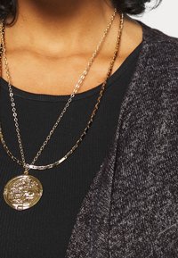 Náhrdelníky ve vrstveném zlatém tónu přes černý žebrovaný top, s kulatým přívěskem s propracovaným vzorem a texturovaný šedý kardigan.
