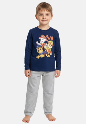 Junger Junge steht barfuß, trägt ein dunkelblaues Langarmshirt mit Cartoons von Welpen und graue Jogginghose, lächelt leicht vor weißem Hintergrund.