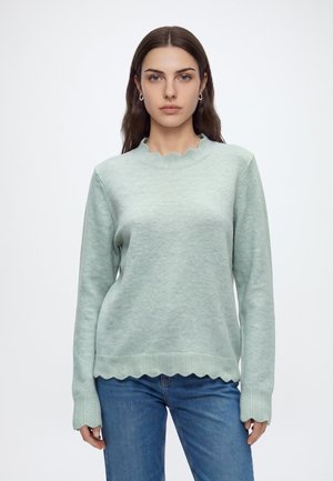 Femme aux longs cheveux bruns portant un pull vert clair texturé avec des bords festonnés et un jean bleu, debout devant un fond uni.