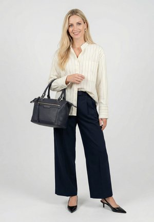 Nainen, jolla on päällään kermansävyinen raidallinen paita, tummansiniset leveälahkeiset housut ja mustat korkokengät, pitää mustaa nahkaista käsilaukkua, seisoo yksiväristä vaaleaa taustaa vasten.