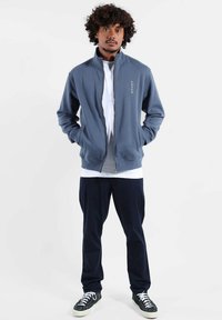 Homme aux cheveux bouclés portant une veste bleue zippée, une chemise blanche, un pantalon foncé et des baskets noires, debout les mains dans les poches.