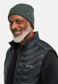 Uomo sorridente con barba grigia che indossa un berretto verde e un gilet imbottito nero Jack Wolfskin sopra una maglietta nera, rivolto leggermente a sinistra.