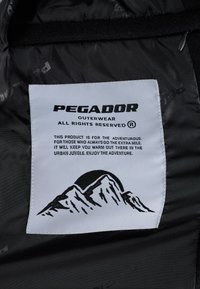 Melnā iekšējā oderējuma ar baltu etiķeti, uz kuras redzams "PEGADOR" logotips. Etiķete ietver kalnu grafiku un tekstu par piedzīvojumu izmantošanu.