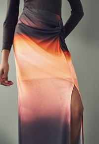 Jupe maxi dégradé orange et noir, avec un tissu texturé, un détail de taille nouée et une fente latérale pour un design supplémentaire.
