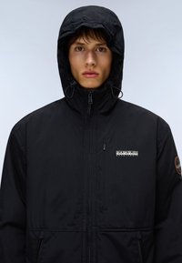 Svart vattentät jacka med dragkedja fram, justerbar huva och logotyp på bröstet. Har slät tyg och elastiska ärmslut.