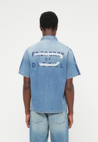 Πουκάμισο από τζιν με κοντά μανίκια, ανοιχτό μπλε χρώμα, με λευκό και ναυτικό μπλε γραφικό τύπωμα στην πλάτη που αναγράφει "PROPERTY OF DIESEL."