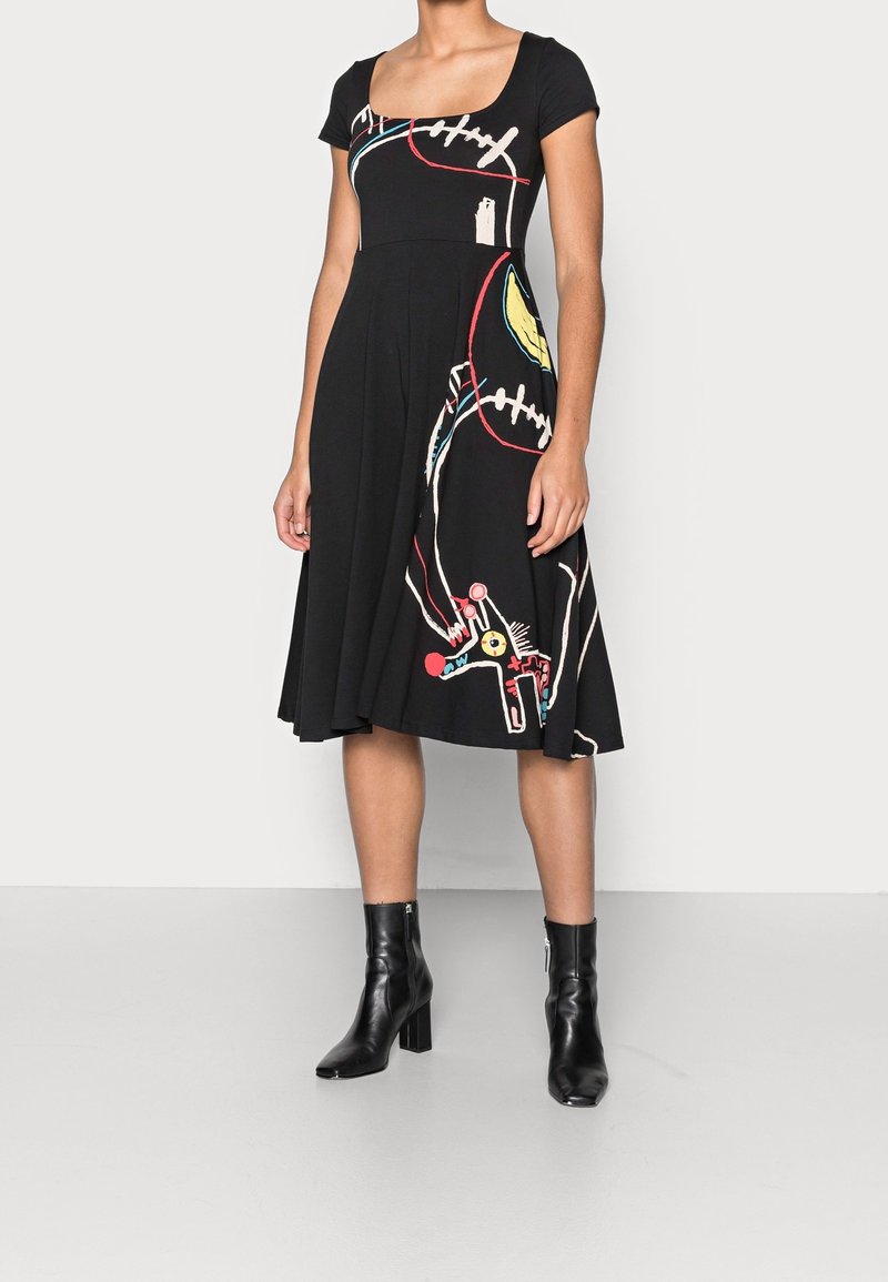 Robe noire à manches courtes avec un décolleté carré, présentant des motifs abstraits colorés et une jupe évasée. Associée à des bottines noires à talons.