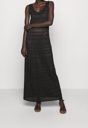 Mujer con un vestido largo negro sin mangas con patrones geométricos de encaje, combinado con sandalias planas negras con tiras.