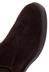 Donkerbruine suede schoen met een afgeronde punt en een dikke donkere zool, die gedetailleerde stikken langs de rand heeft.