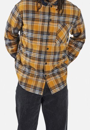 Personne portant une chemise à carreaux jaune et bleu marine boutonnée avec un pantalon noir, mains dans le dos, cadrée aux épaules et aux cuisses.