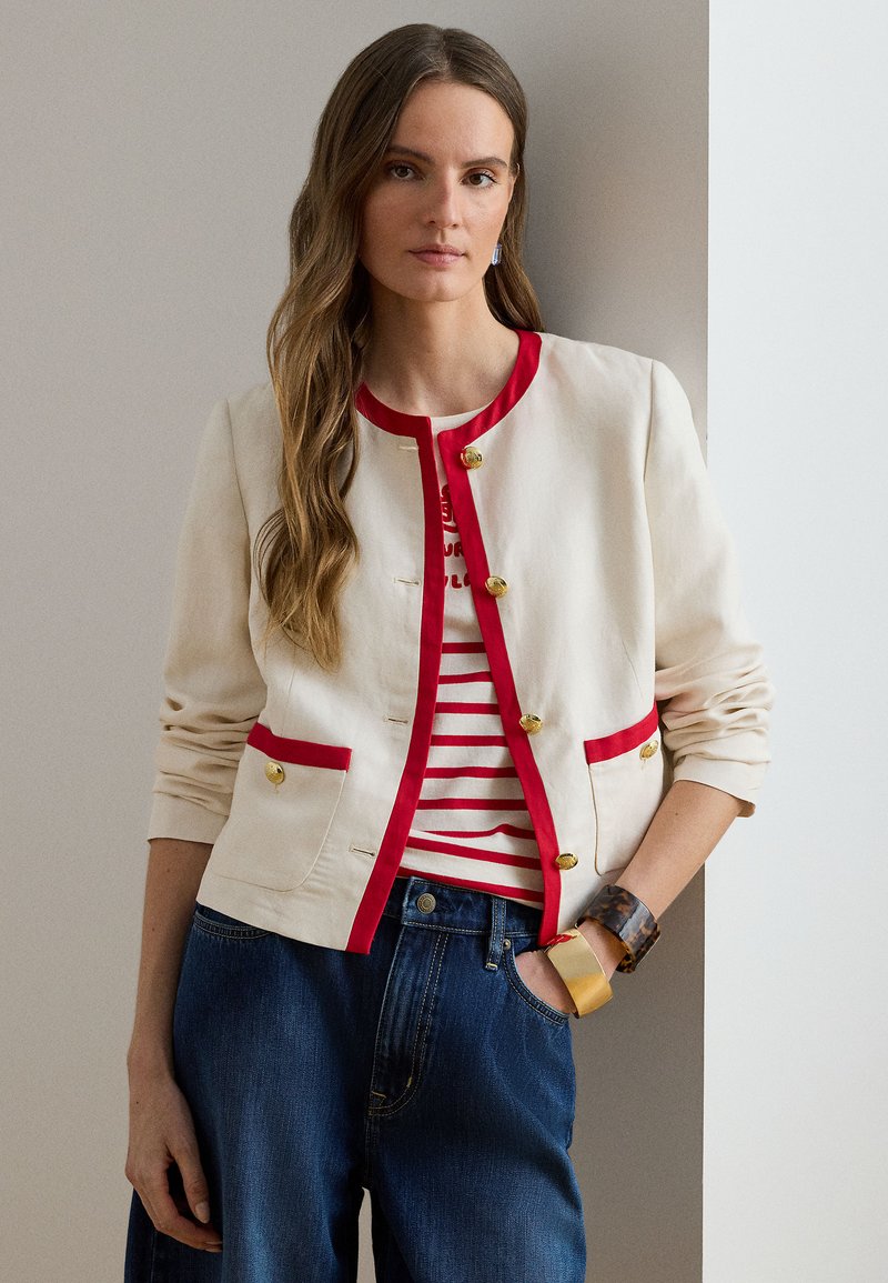 Εκρού μπουφάν με κόκκινες επενδύσεις, χρυσά κουμπιά και τσέπες. Φορεμένο πάνω από ριγέ μπλούζα και συνδυασμένο με μπλε τζιν. Λεία υφή και casual σχεδιασμός.