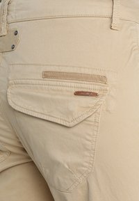 Beżowe spodnie z zapinaną na guzik tylną kieszenią, z małą prostokątną skórzaną metką z tekstem marki.