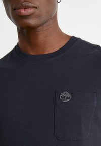 Tricou scurt de culoare bleumarin, cu guler rotund și un buzunar pe piept la stânga. Buzunarul are un logo brodat cu un copac. Textură moale.