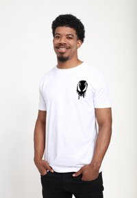 Bílá bavlněná tričko s krátkým rukávem, na kterém je černý grafický design na levém prsu. Model má na sobě černé kalhoty a stojí se založenýma rukama v kapsách.