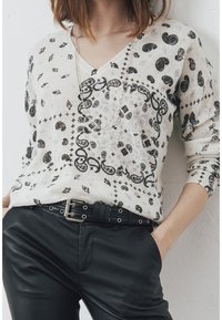 Pull crème avec un col en V, orné de motifs paisley noirs et géométriques, associé à un pantalon en cuir noir et une ceinture avec des accessoires en argent.