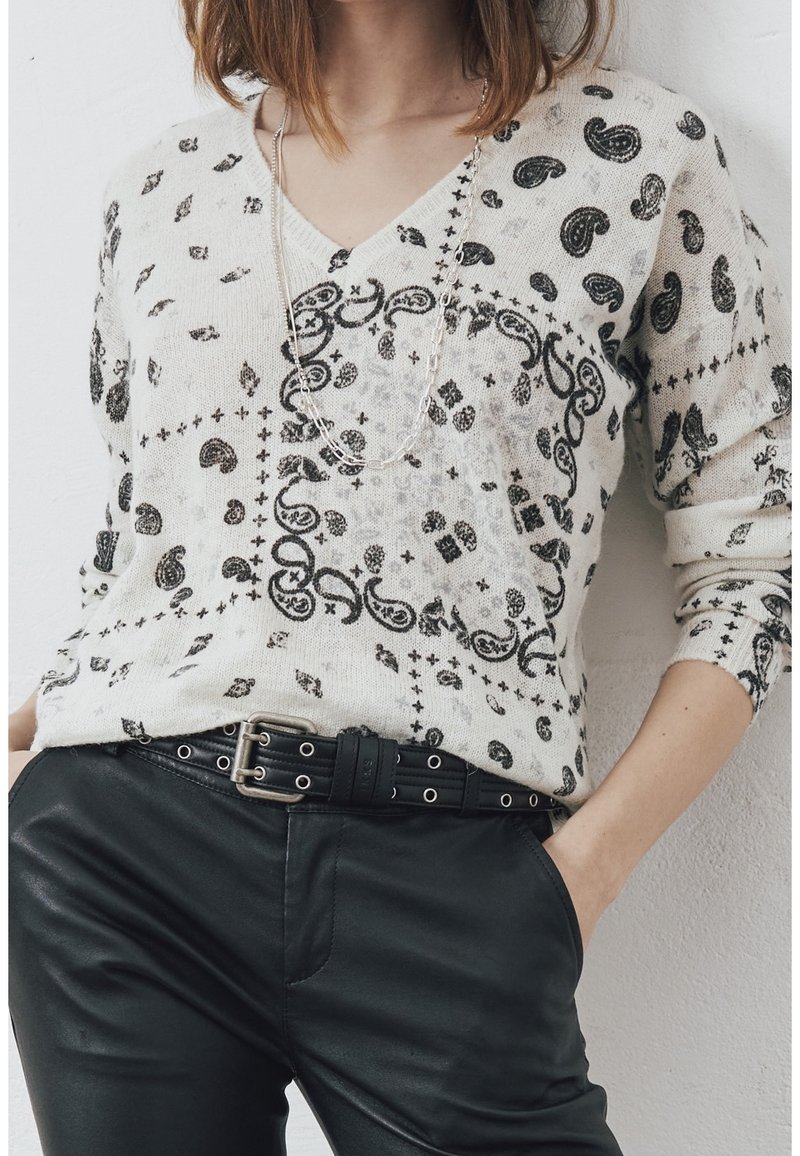 Maglione crema con scollatura a V, caratterizzato da intricate fantasie paisley nere e geometriche, abbinato a pantaloni in pelle nera e una cintura con accessori in argento.