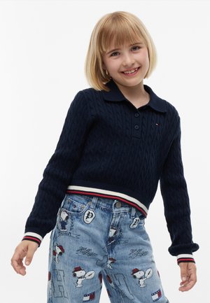 Dziecko z blond fryzurą na boba uśmiecha się, nosząc granatowy sweter w warkocze i jasnoniebieskie dżinsy z nadrukami z kreskówki Snoopy.