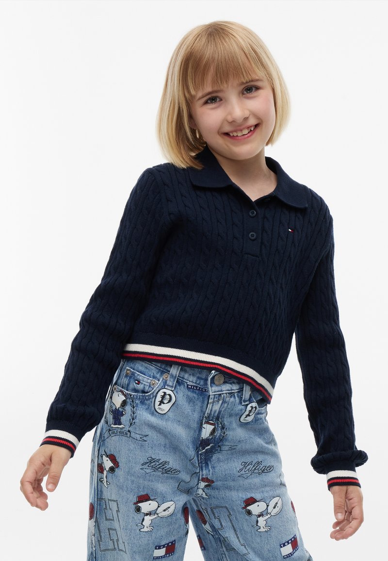 Παιδί με ξανθό καρέ χαμογελά, φοράει μπλε πουλόβερ με πλεκτό σχέδιο και ανοιχτό μπλε τζιν με εκτυπώσεις καρτούν Snoopy.