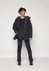 Zwarte waterdichte parka met verstelbare capuchon, ritszakken; gecombineerd met zwarte broek met knieverstevigingen en rode wandelschoenen.