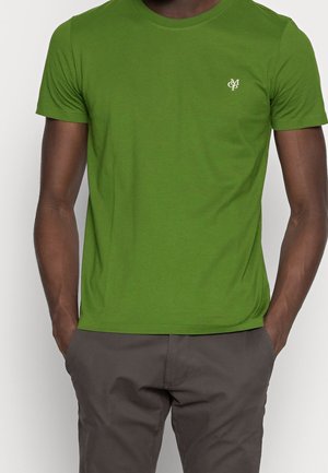 Homme portant un t-shirt ajusté vert vif à manches courtes avec un petit logo blanc sur la poitrine, assorti à un pantalon gris foncé, les mains dans les poches.