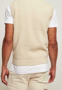 Beżowy dzianinowy bezrękawnik o prążkowanej strukturze, z krótkimi rękawami i prostym wykończeniem, noszony na białej koszulce, w zestawieniu z jasnymi spodniami.