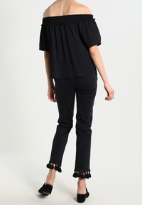 Haut noir épaules dénudées avec encolure smockée, associé à un jean noir avec ourlet effiloché. Le mannequin porte des chaussures plates noires.